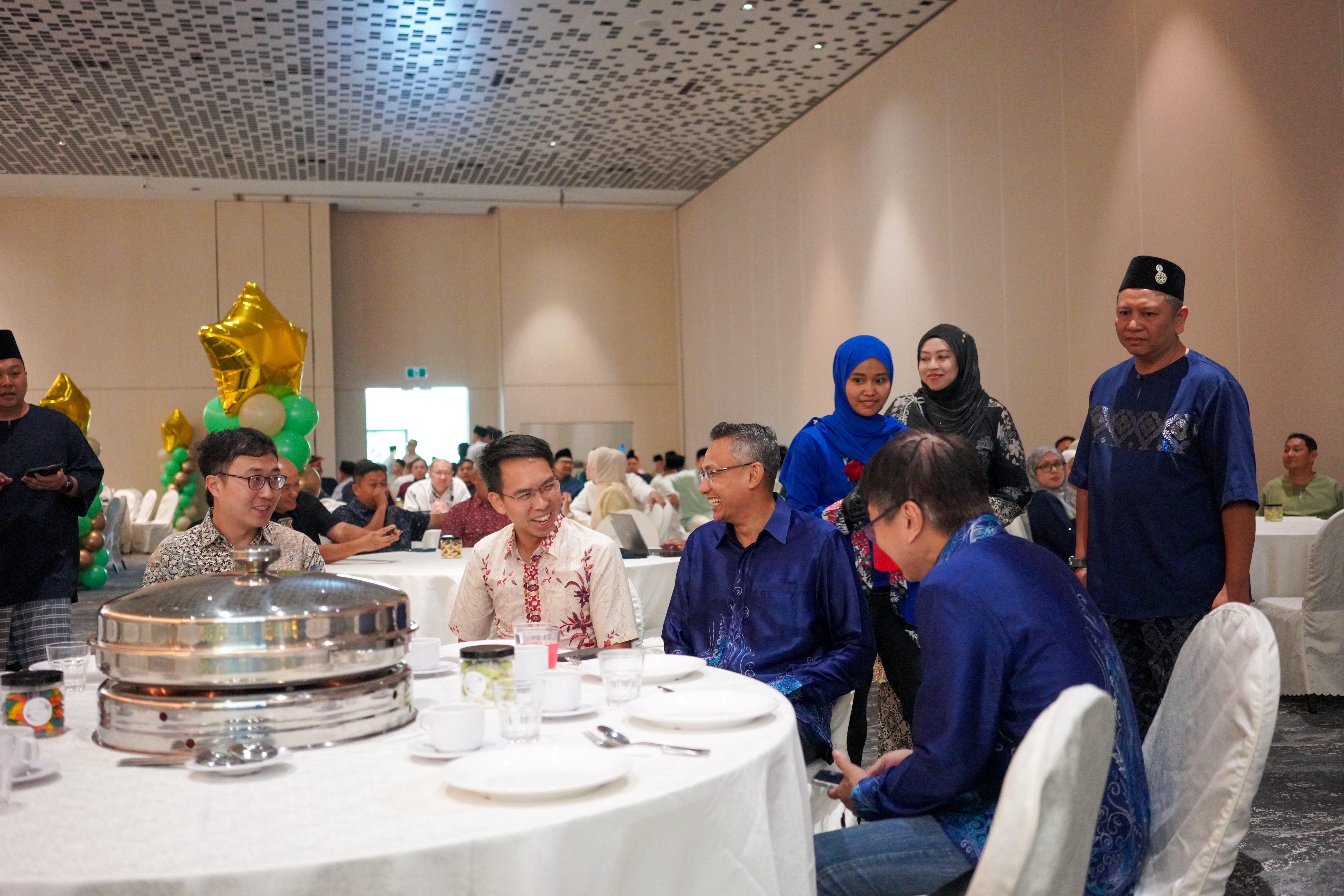 Attendees in traditional attire chatting and smiling around a dining table, with other guests seated throughout the banquet hall.